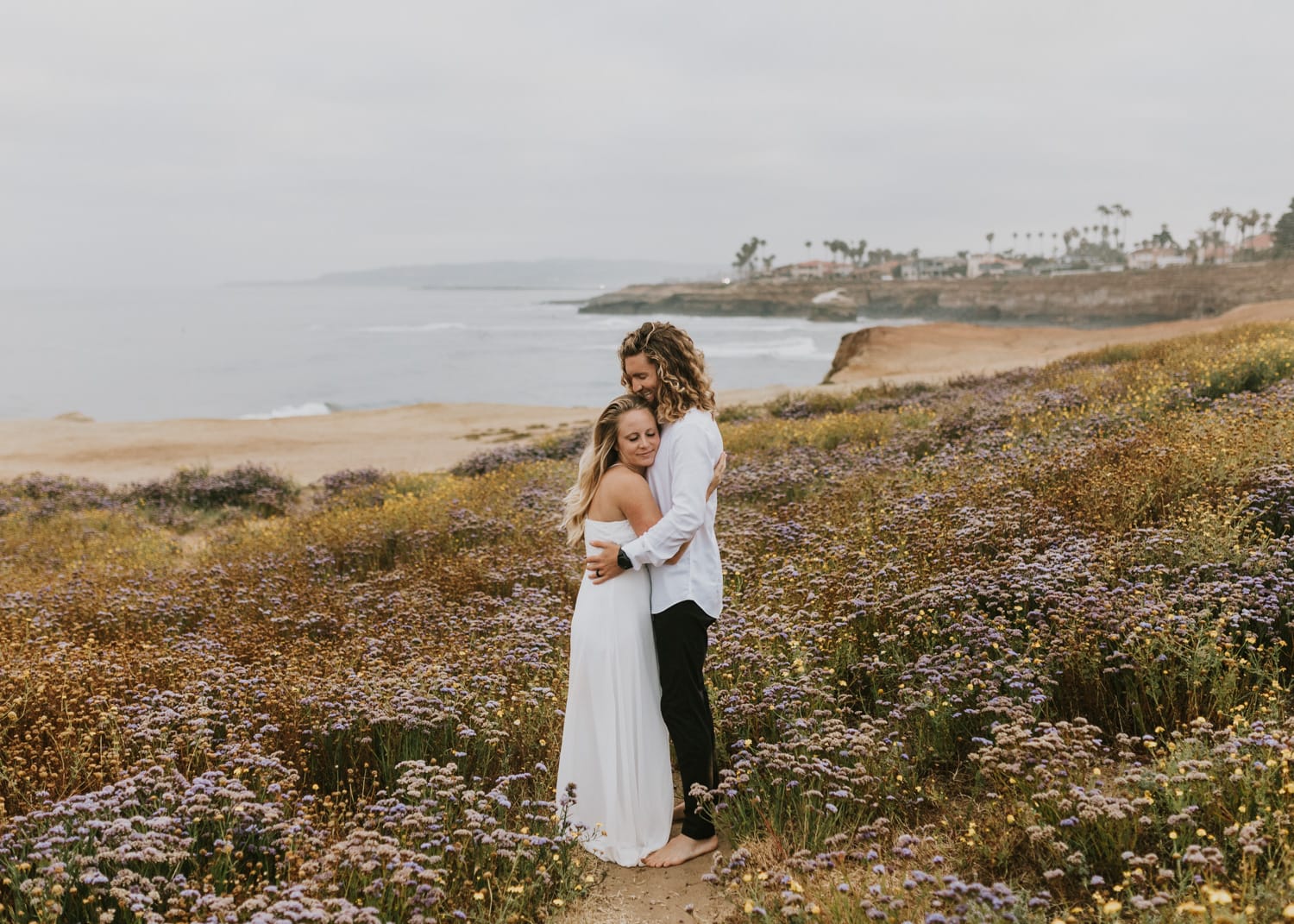 A bride and groom hugging in some wildflowers at Sunset Cliffs Natural Park in San Diego, California during their sunrise elopement.