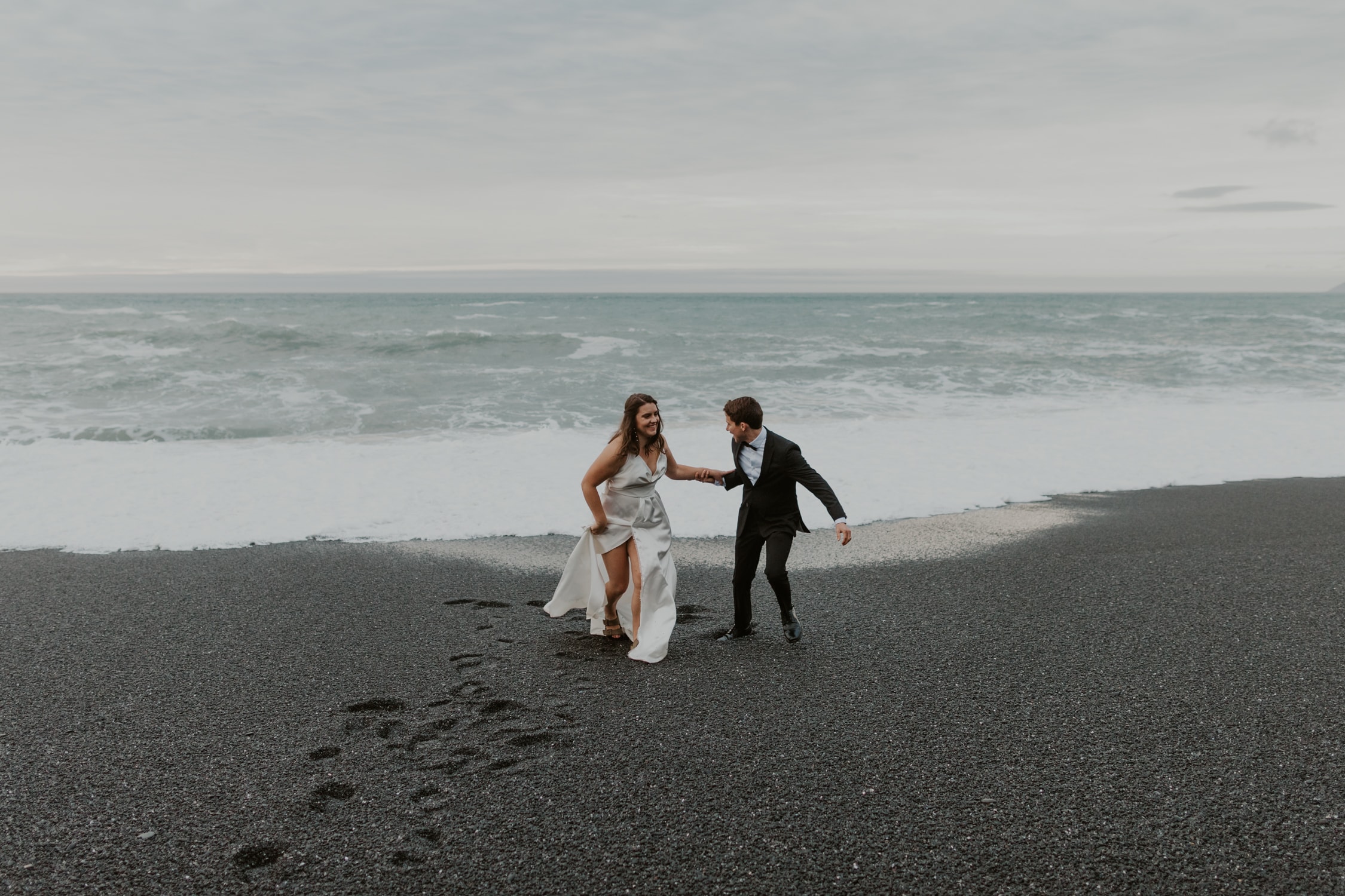 A couple in wedding attire holding hands and running towards the camera on a black sand beach in Northern California.