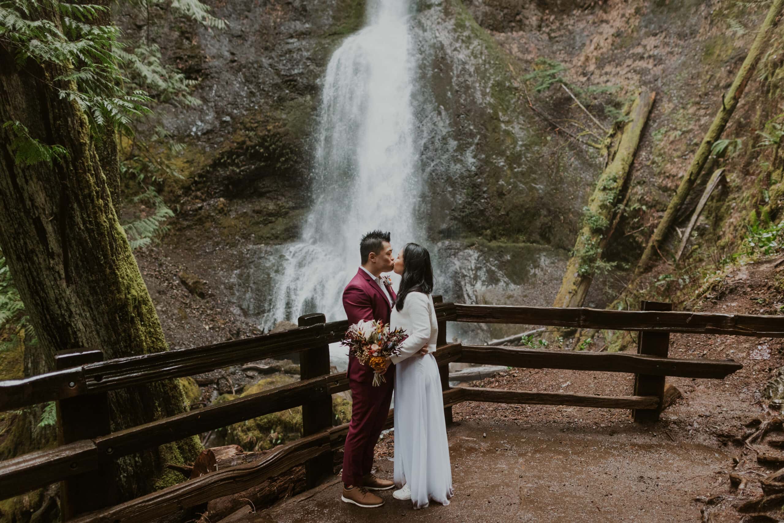 A couple in wedding attire kissing in front of Marymere Falls.