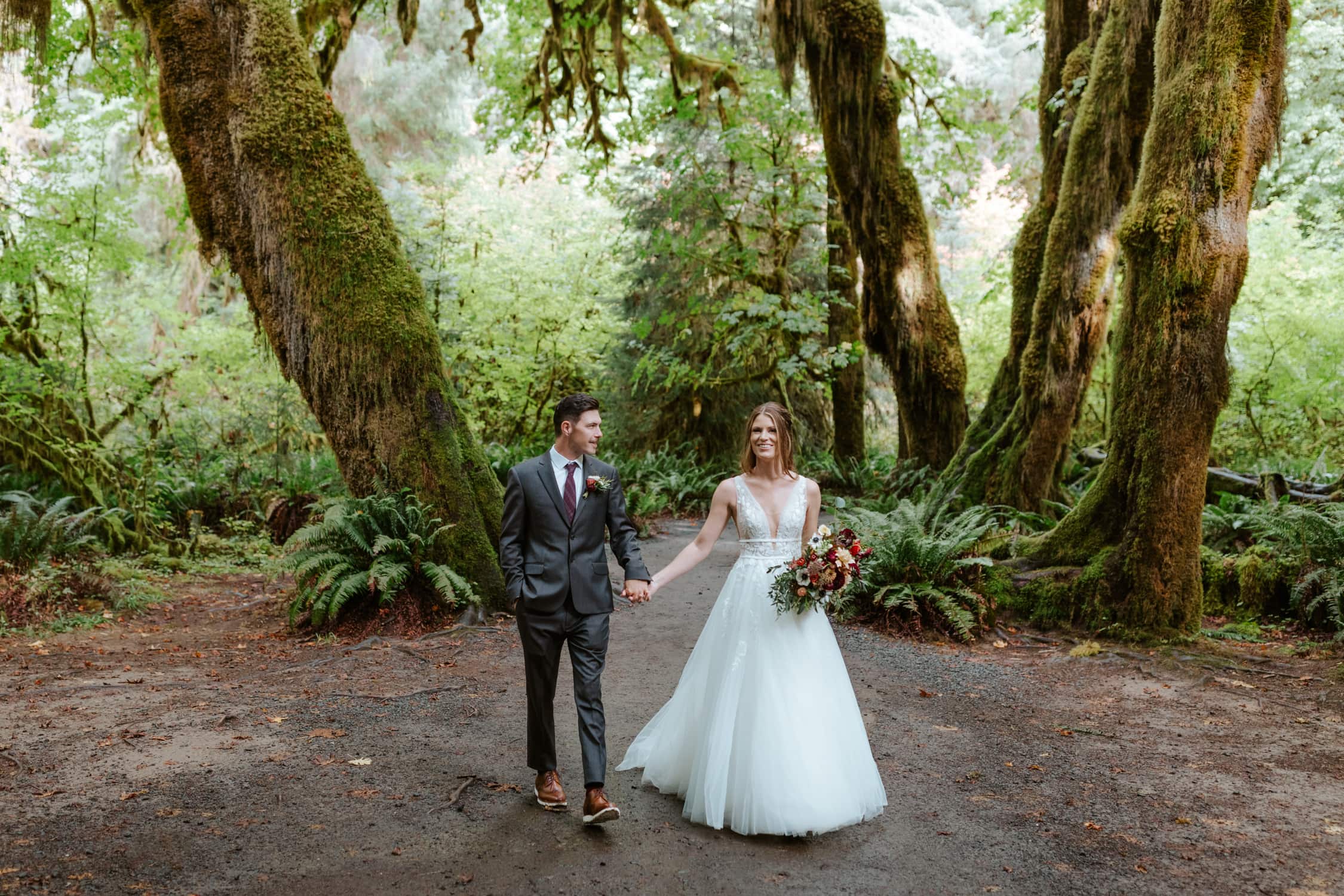A couple in wedding attire holding hands and walking towards the camera in the Hoh Rainforest.