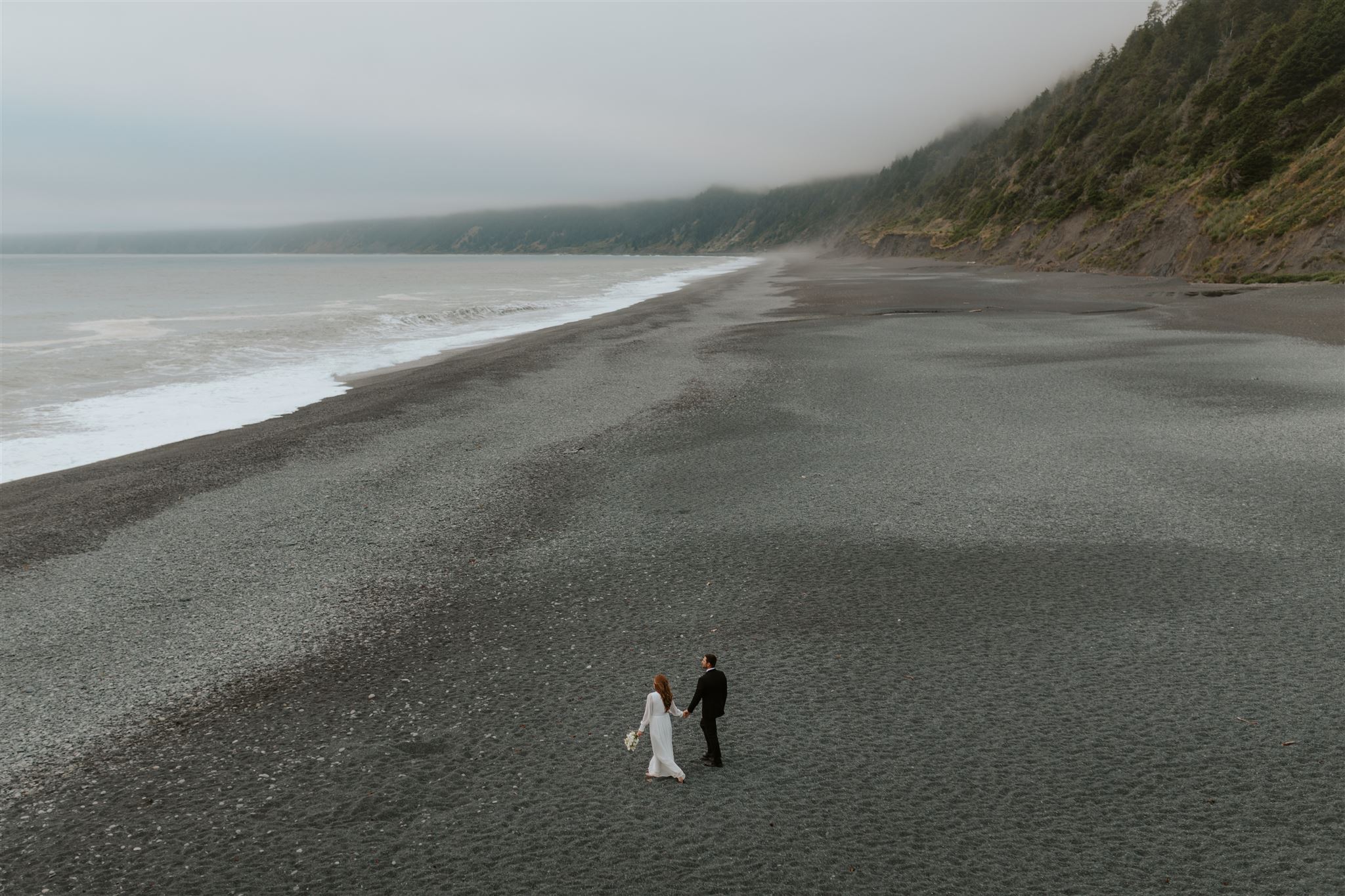 A couple in wedding attire holding hands and walking along a black sand beach in Northern California.