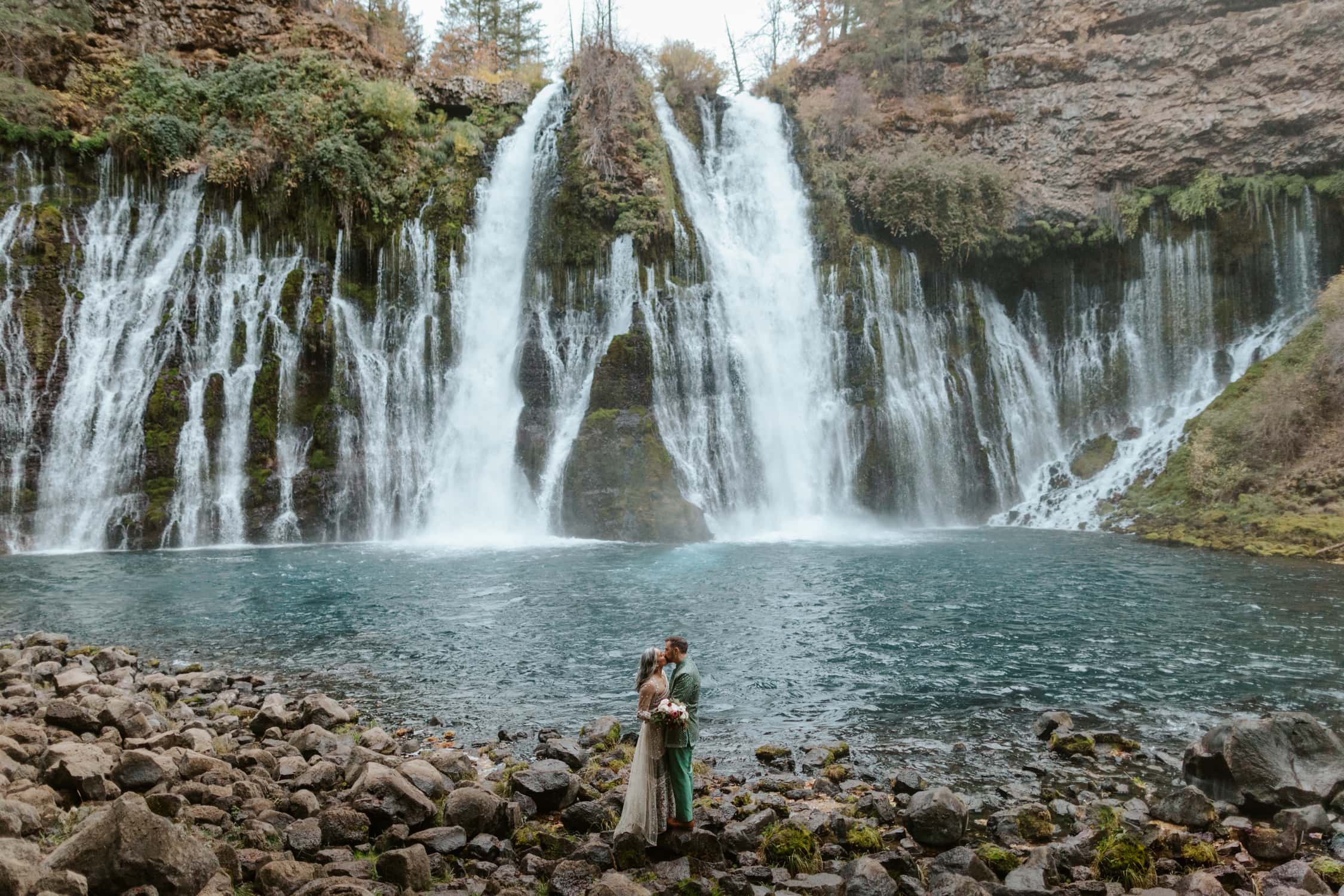 A couple in Indian wedding attire kissing each other in front of Burney Falls in Northern California.