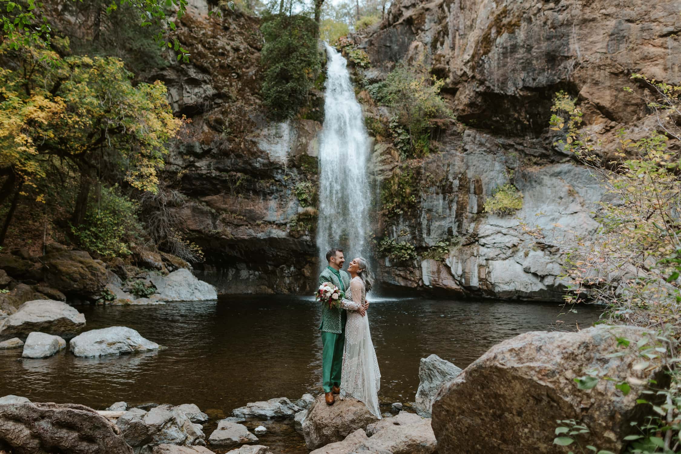 A couple in Indian wedding attire facing each other and laughing in front of a waterfall in Northern California.