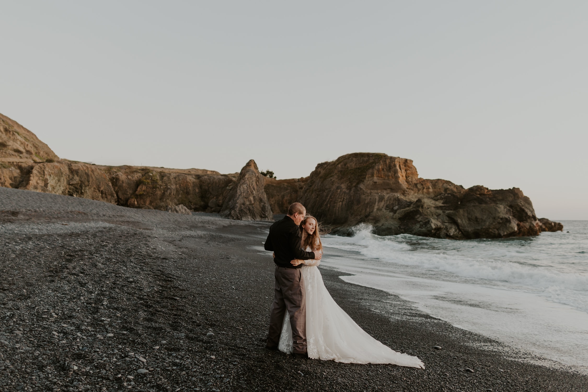 A couple in wedding attire hugging and looking out at the ocean on a black sand beach in Northern California.