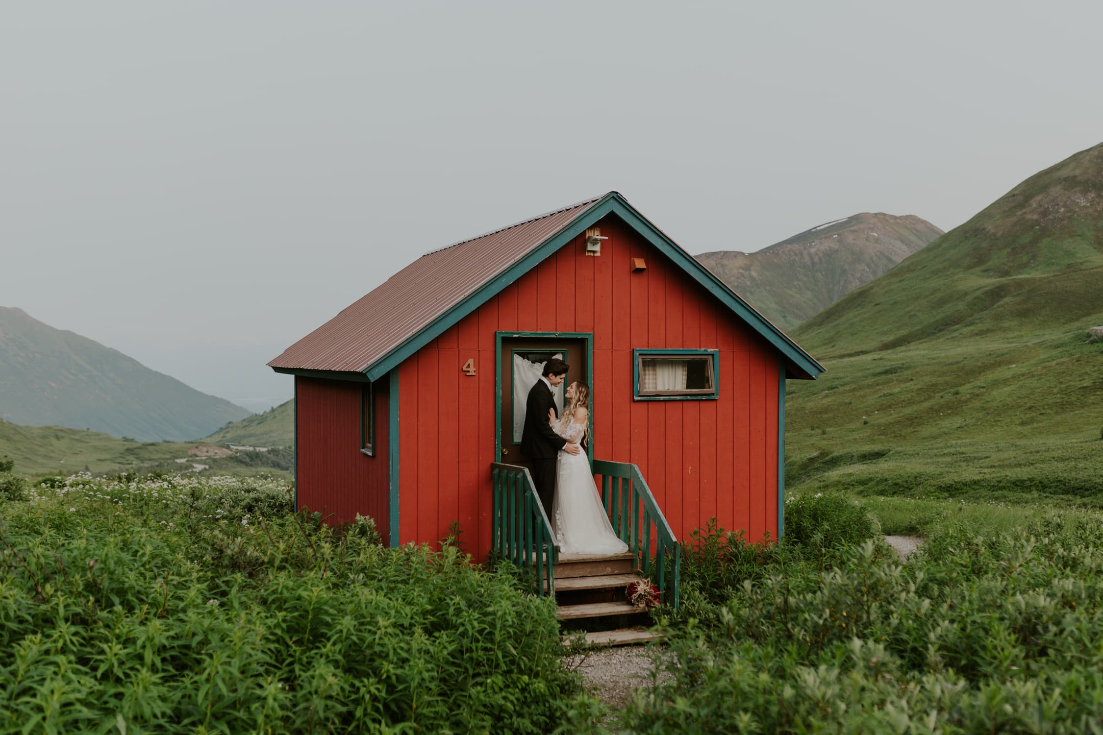 A bride and groom looking up at each other in front of a red cabin in Alaska.