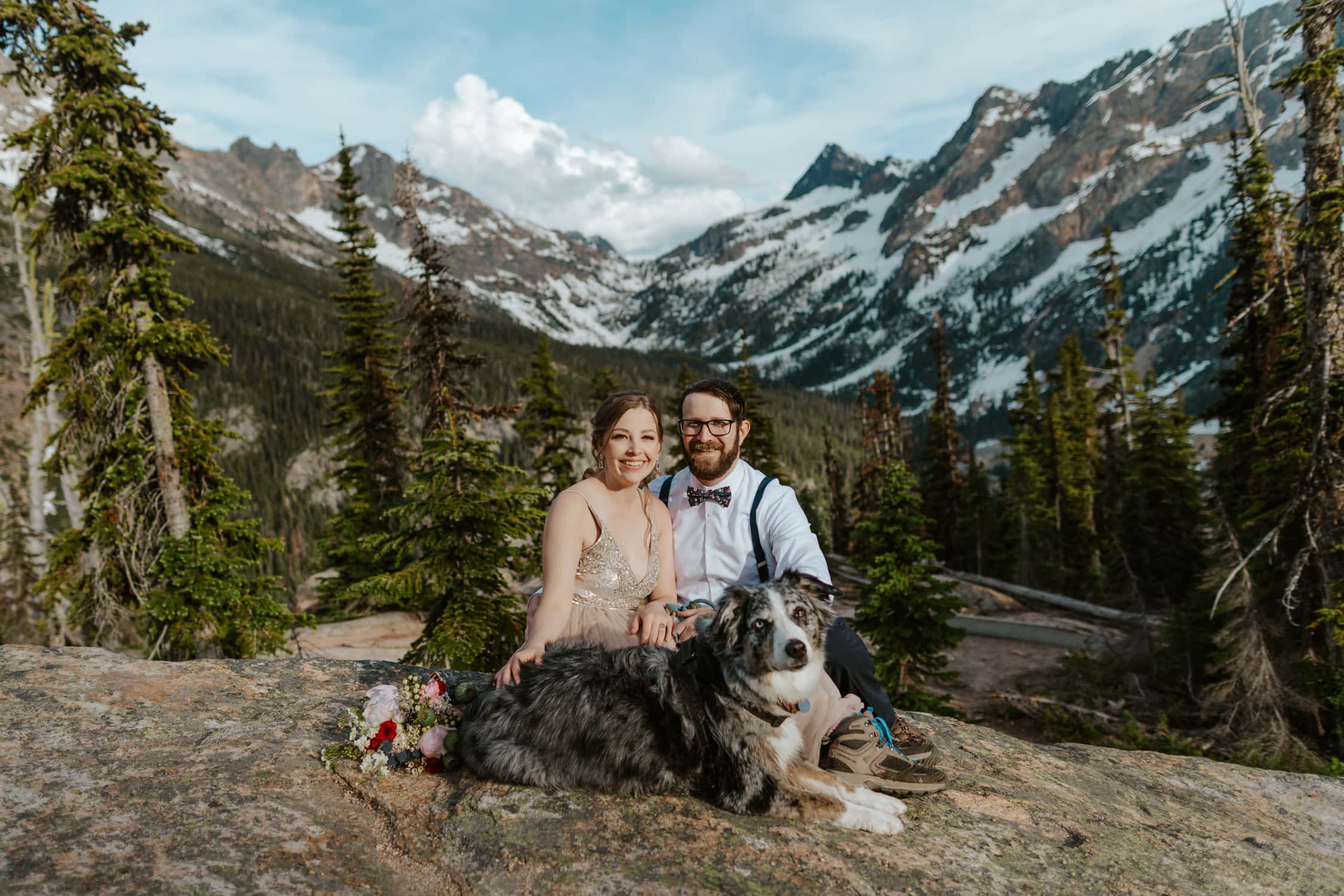 A couple in wedding attire and their Australian Shepard smiling at the camera at Washington Pass Overlook.
