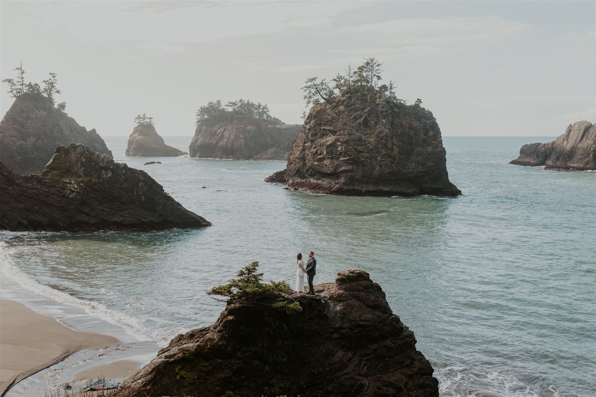 A bride and groom holding hands on top of a rock on the beach.