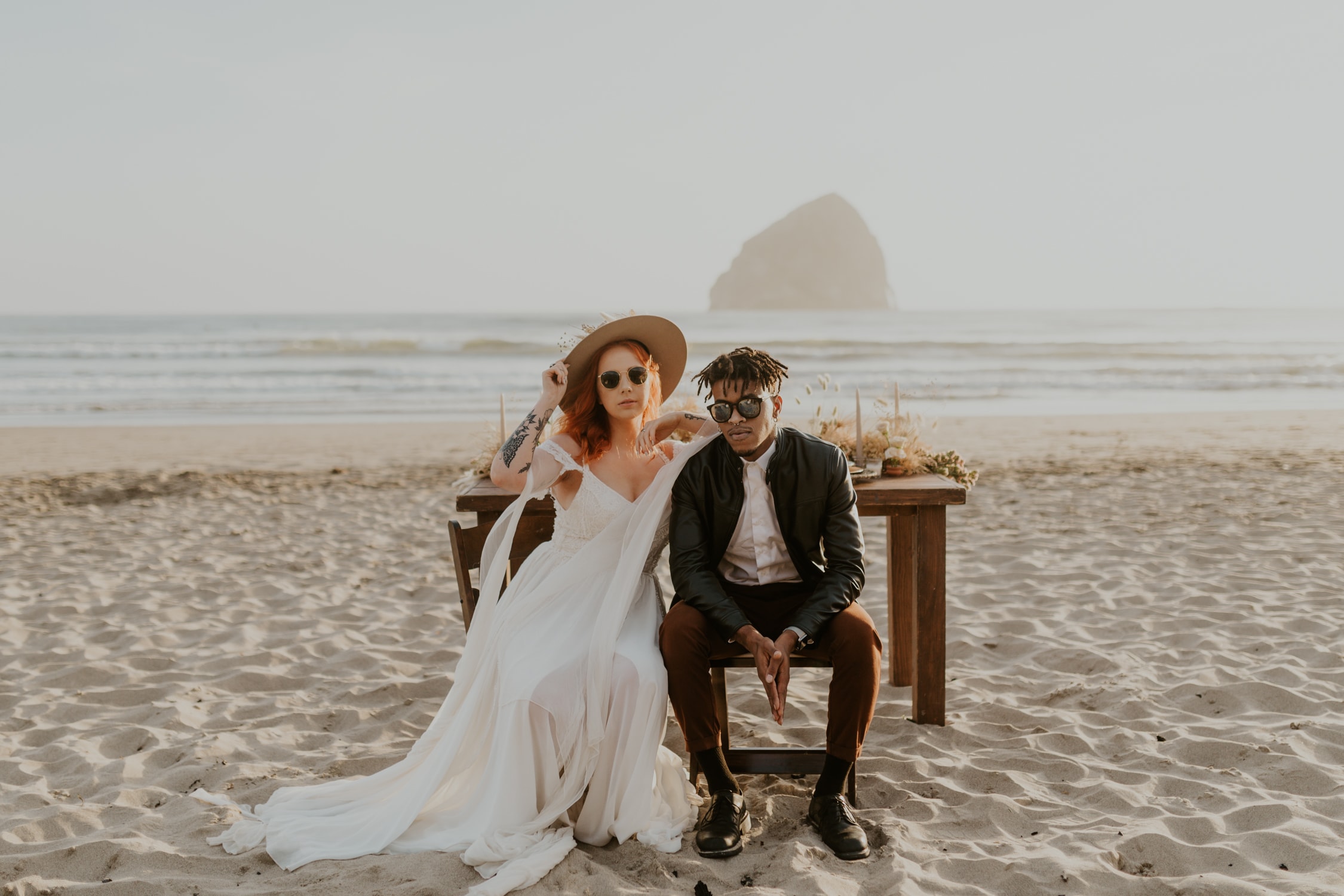 A bride and groom looking at the camera and sitting on chairs on Cannon Beach.