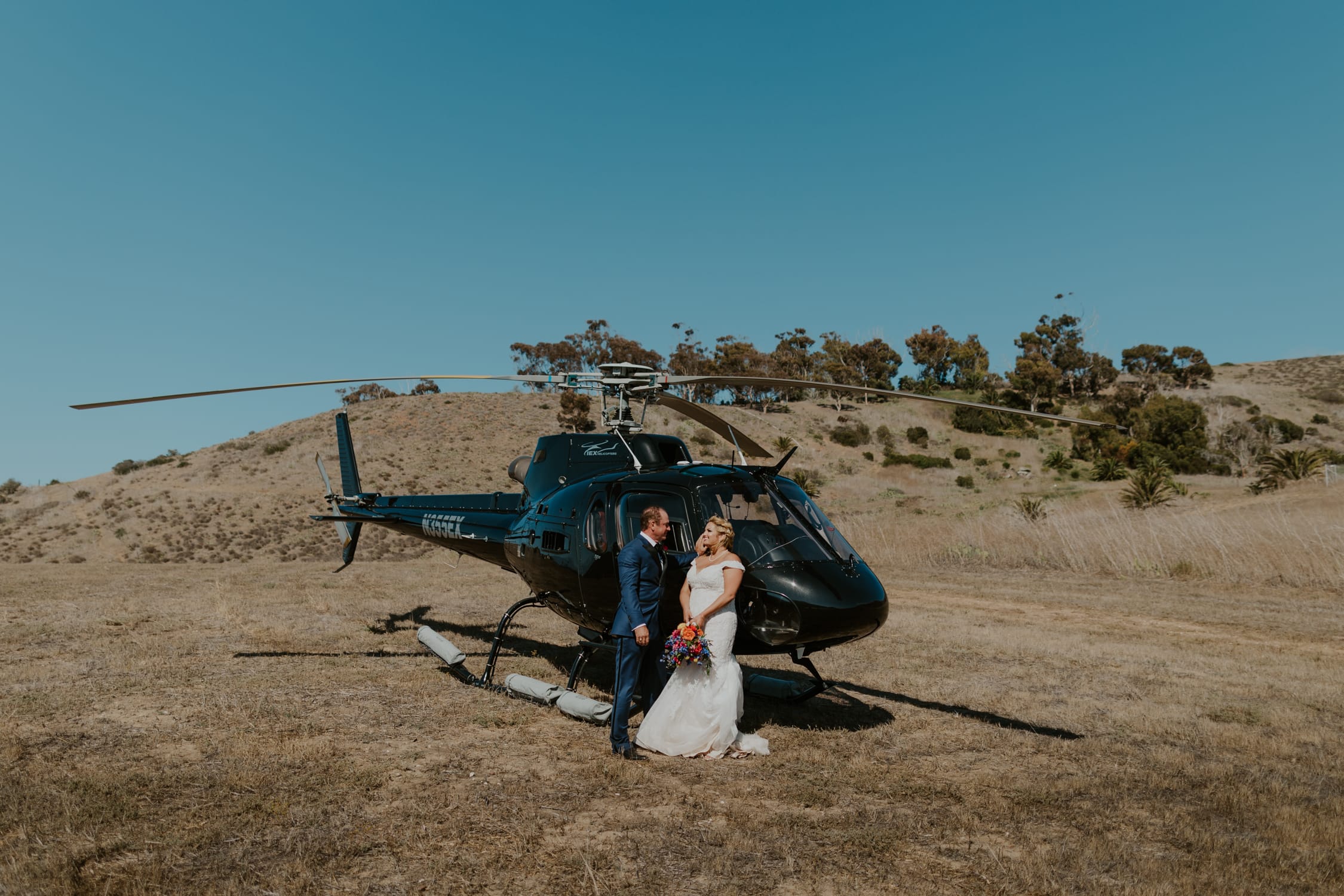 A bride and groom standing on the side of a helicopter on Catalina Island.