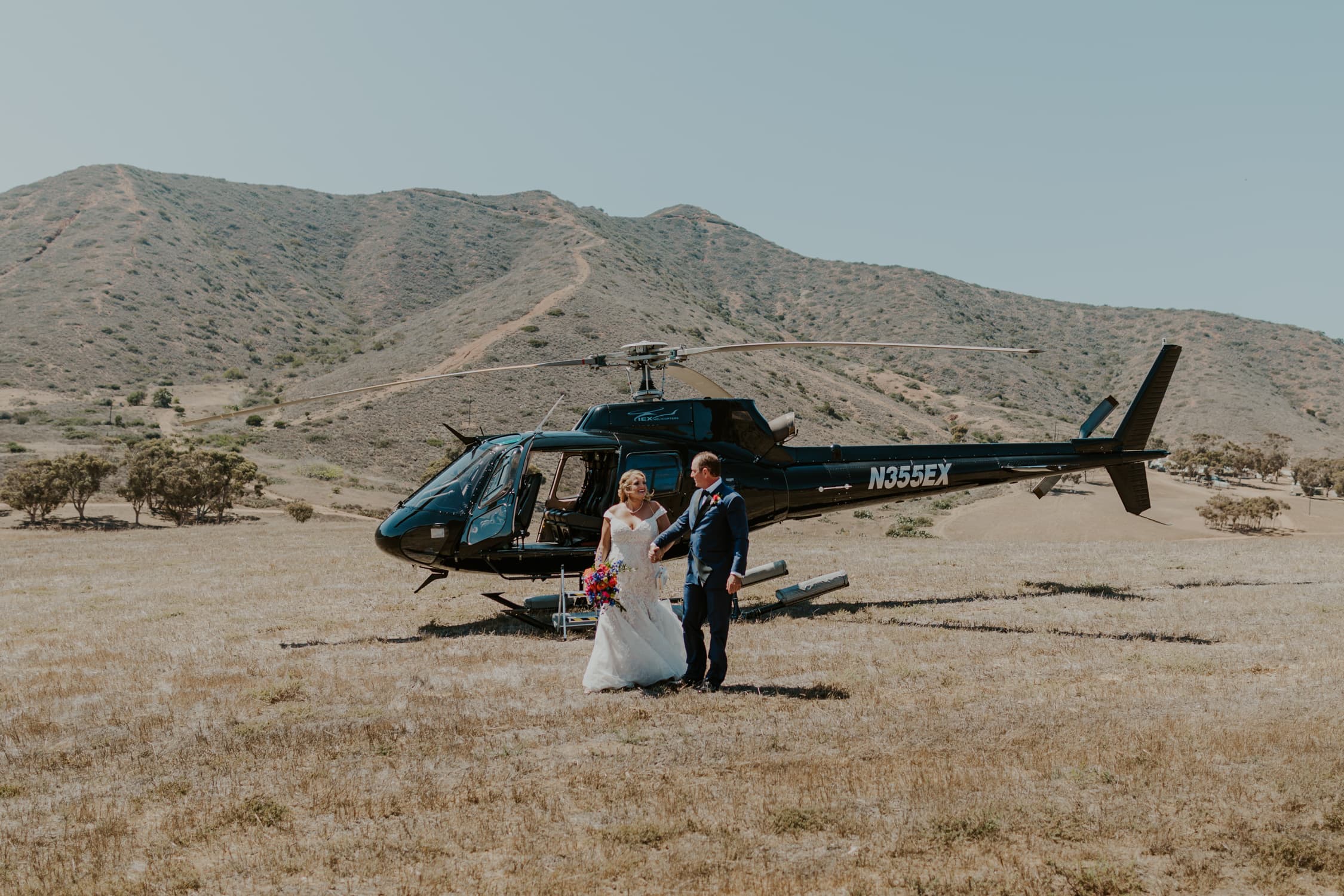 A bride and groom holding hands and walking in front of a helicopter.