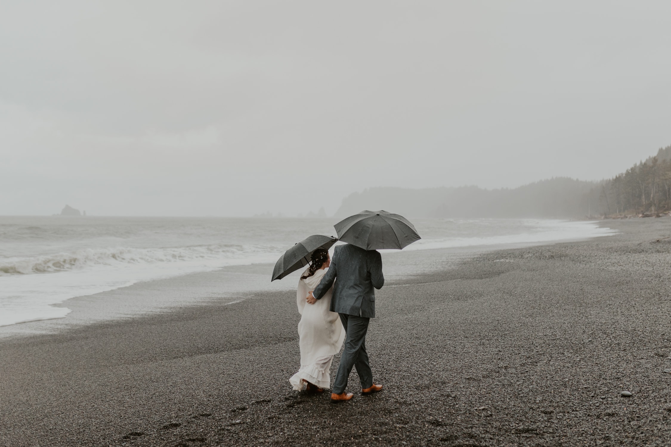 A bride and groom holding umbrellas and walking along Rialto Beach in Olympic National Park.