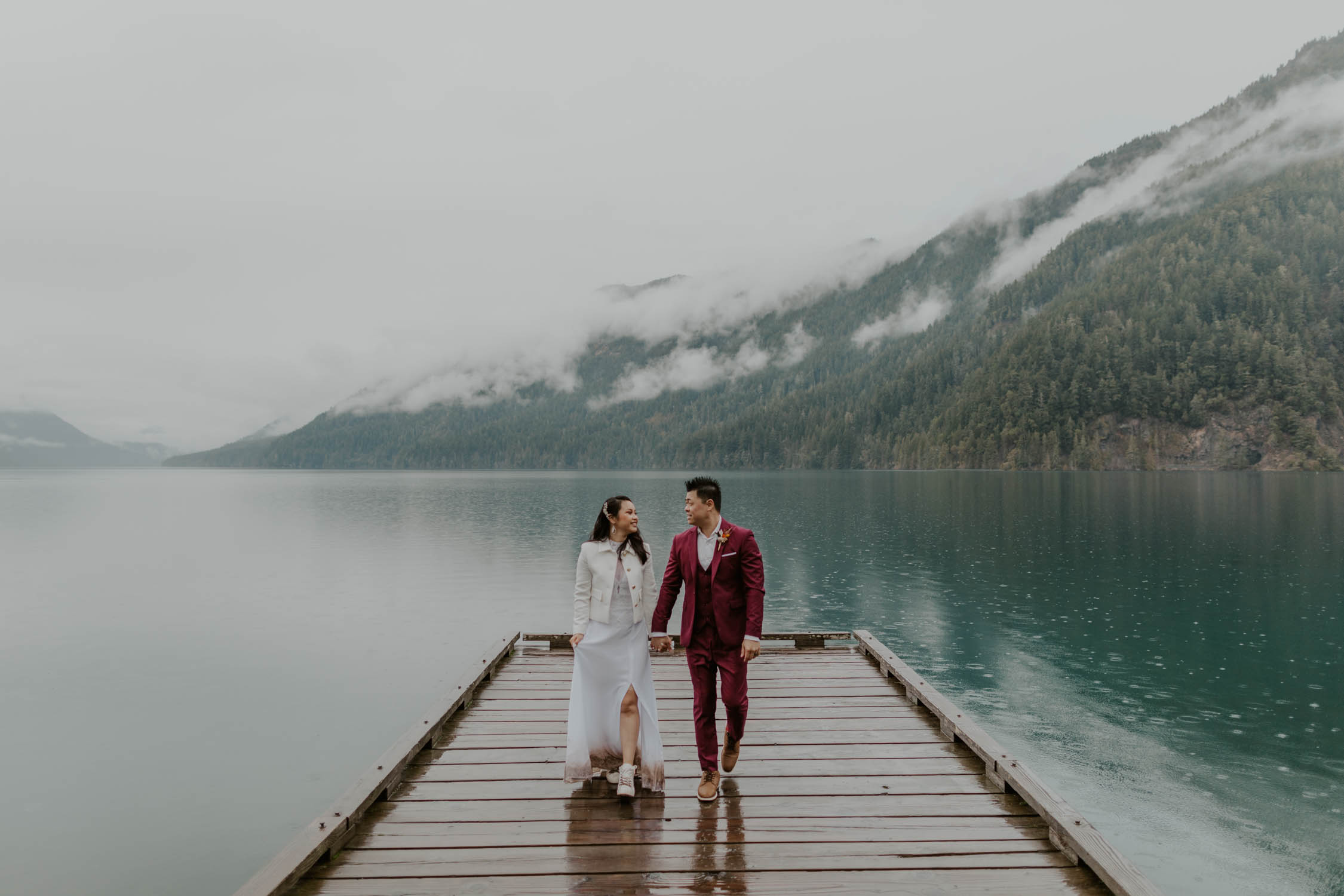 A bride and groom holding hands and walking towards the camera on the dock of Lake Crescent in Olympic National Park on their elopement day.