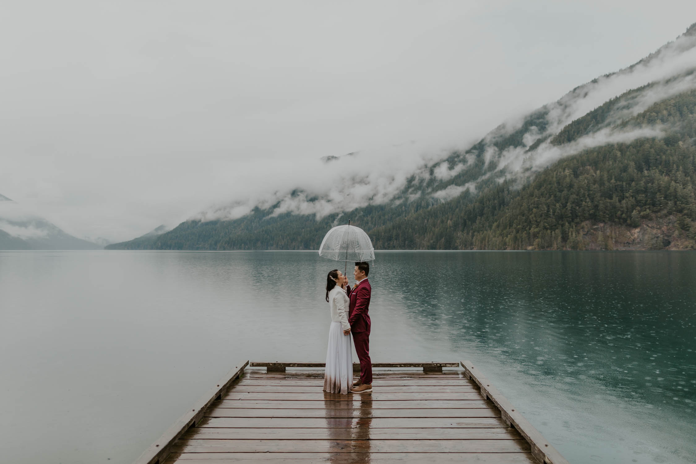 A bride and groom facing each other, holding hands, and holding an umbrella on the dock of Lake Crescent in Olympic National Park on their elopement day.