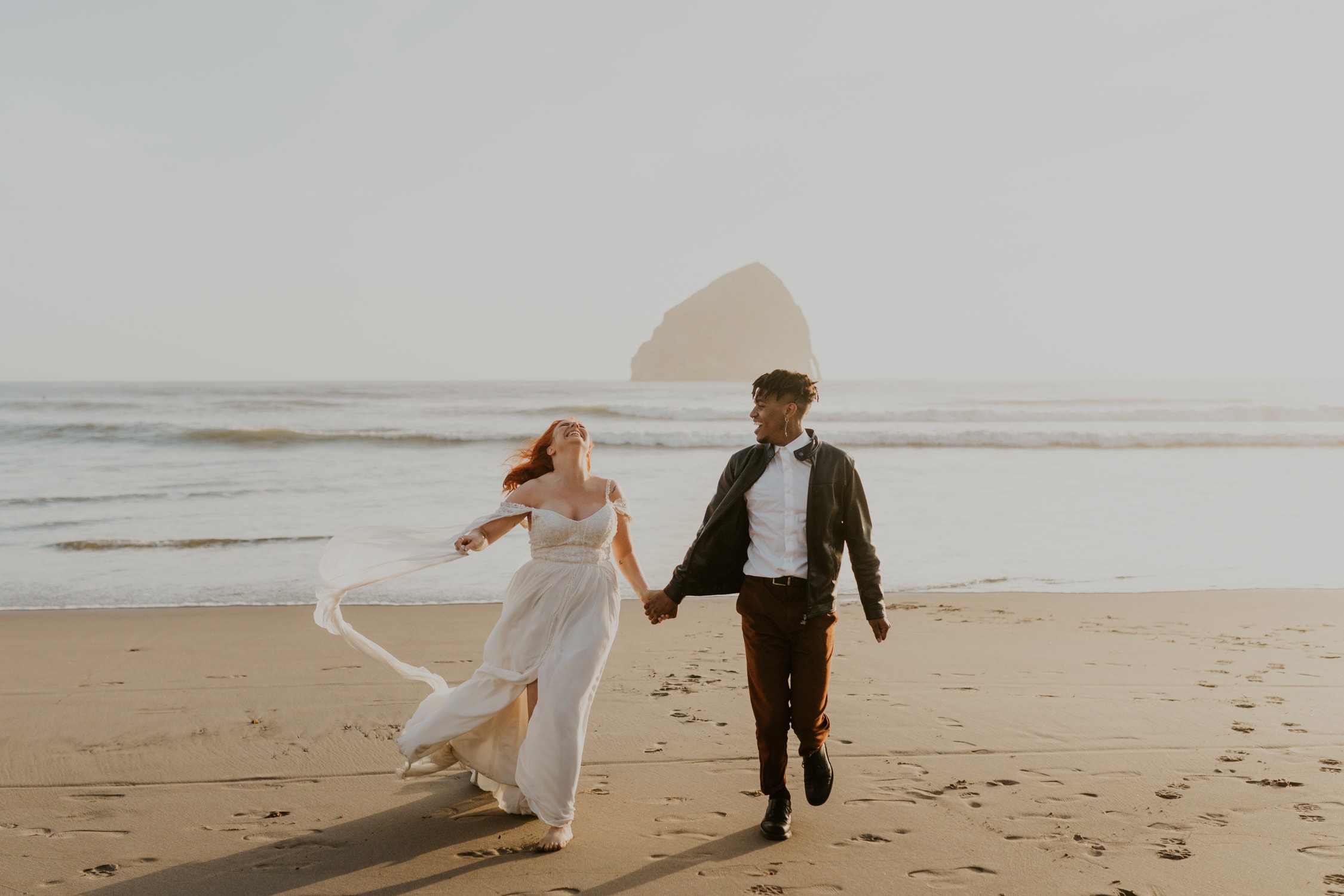 A bride and groom holding hands and running towards the camera on Cape Kiwanda in Oregon.
