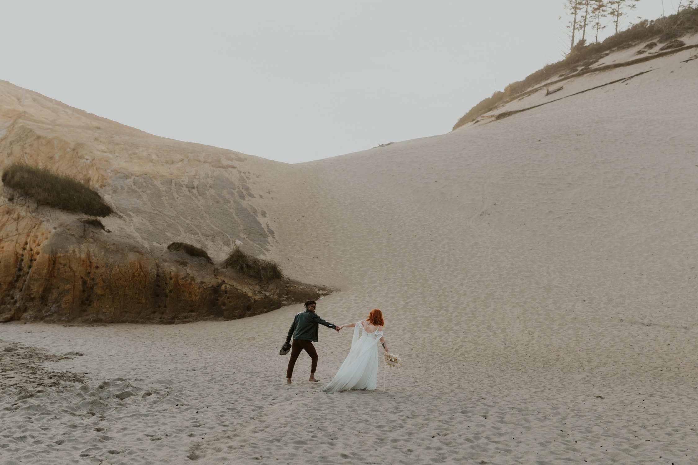 A bride and groom holding hands and walking on Cape Kiwanda in Oregon.