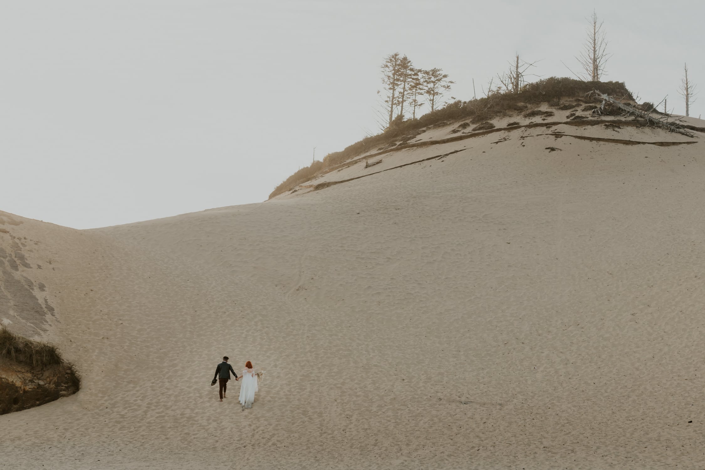 A bride and groom holding hands and walking on Cape Kiwanda in Oregon.