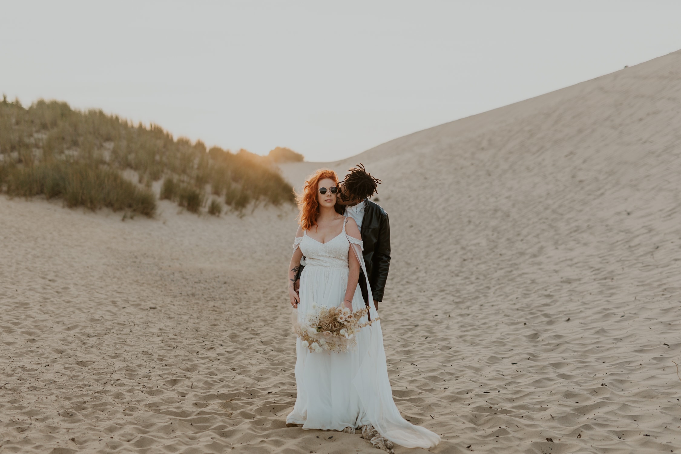 A bride wearing sunglasses looking at the camera while the groom gives her a hug on Cape Kiwanda on Oregon.