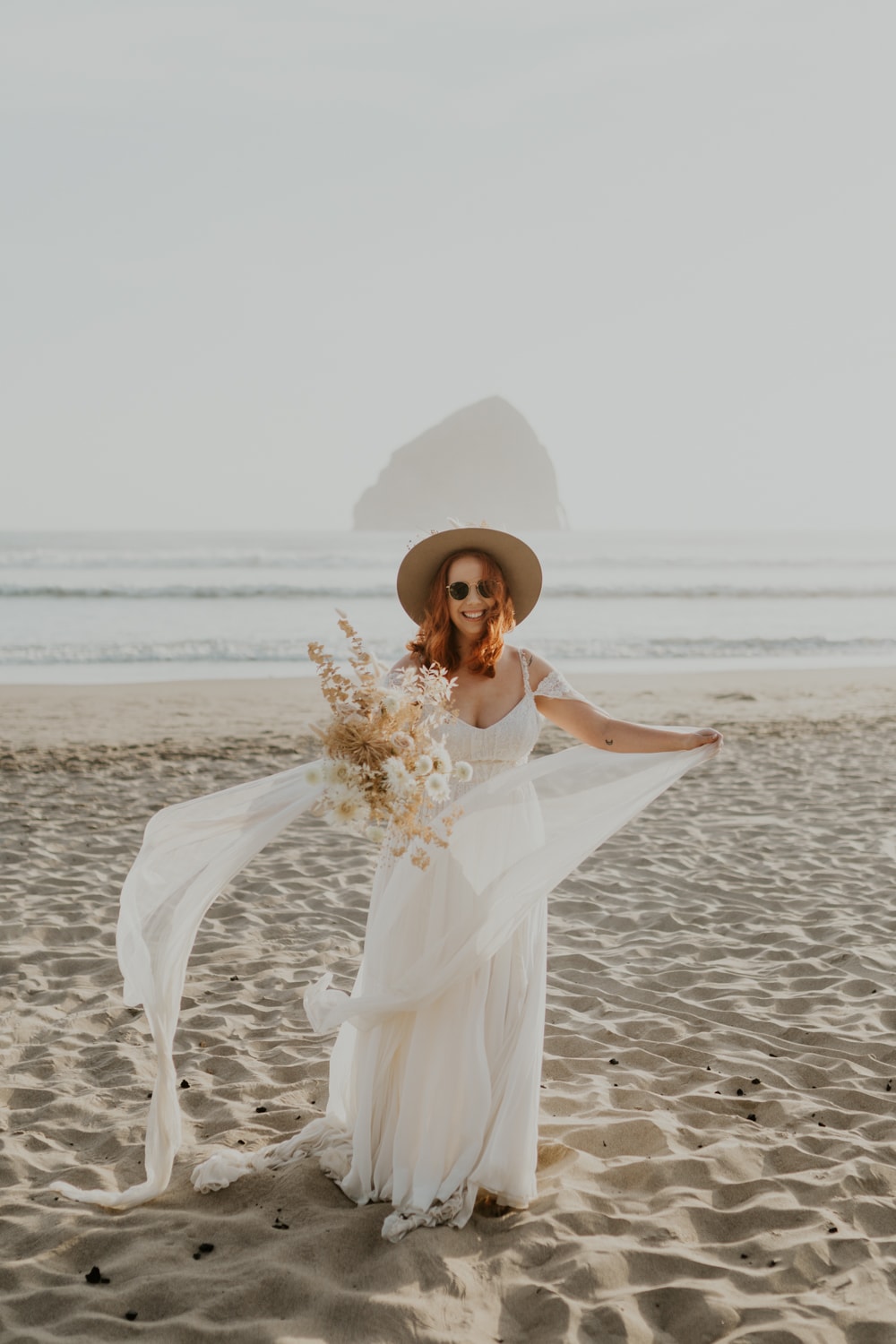 A bride wearing a hat and sunglasses looking at the camera on Cape Kiwanda on Oregon.