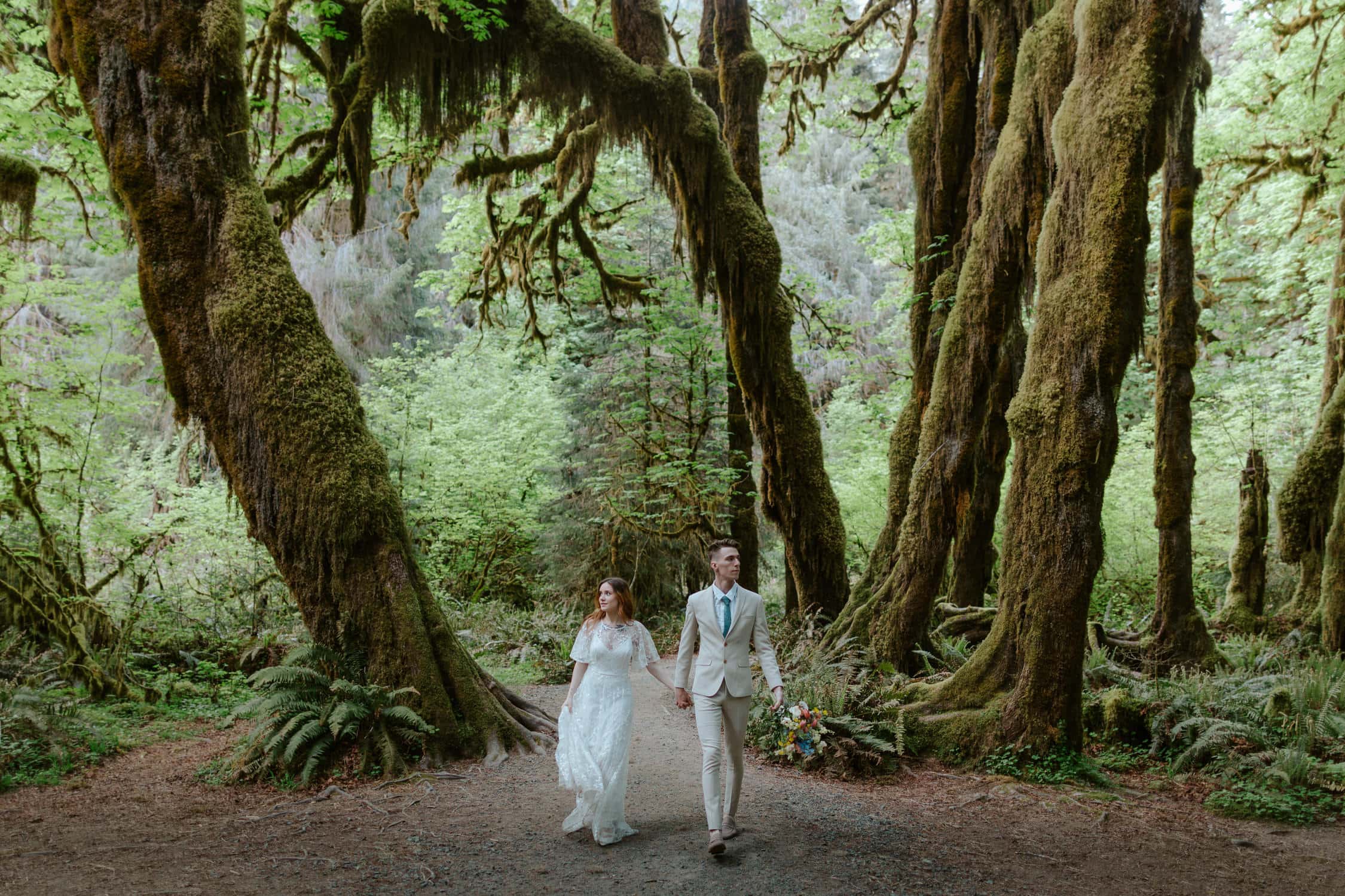 A bride and groom holding hands and walking towards the camera in the Hoh Rainforest on their elopement day.