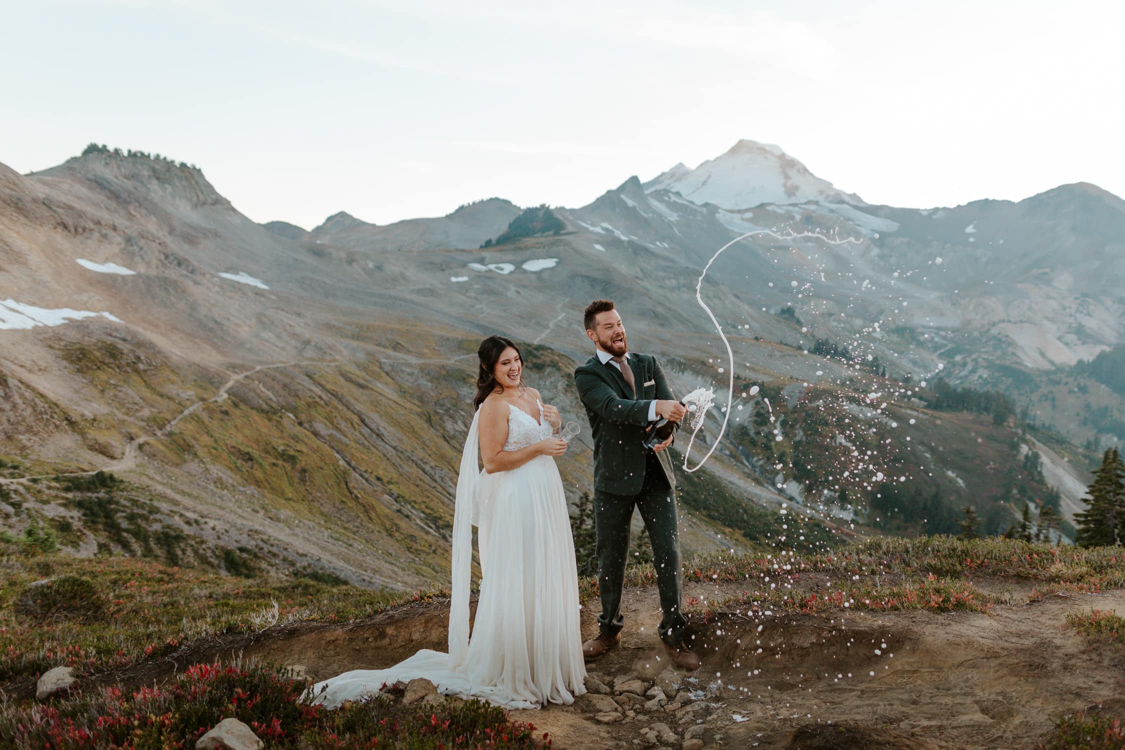 A bride and groom laughing as the groom pops champagne in front of Mt. Baker in Washington.