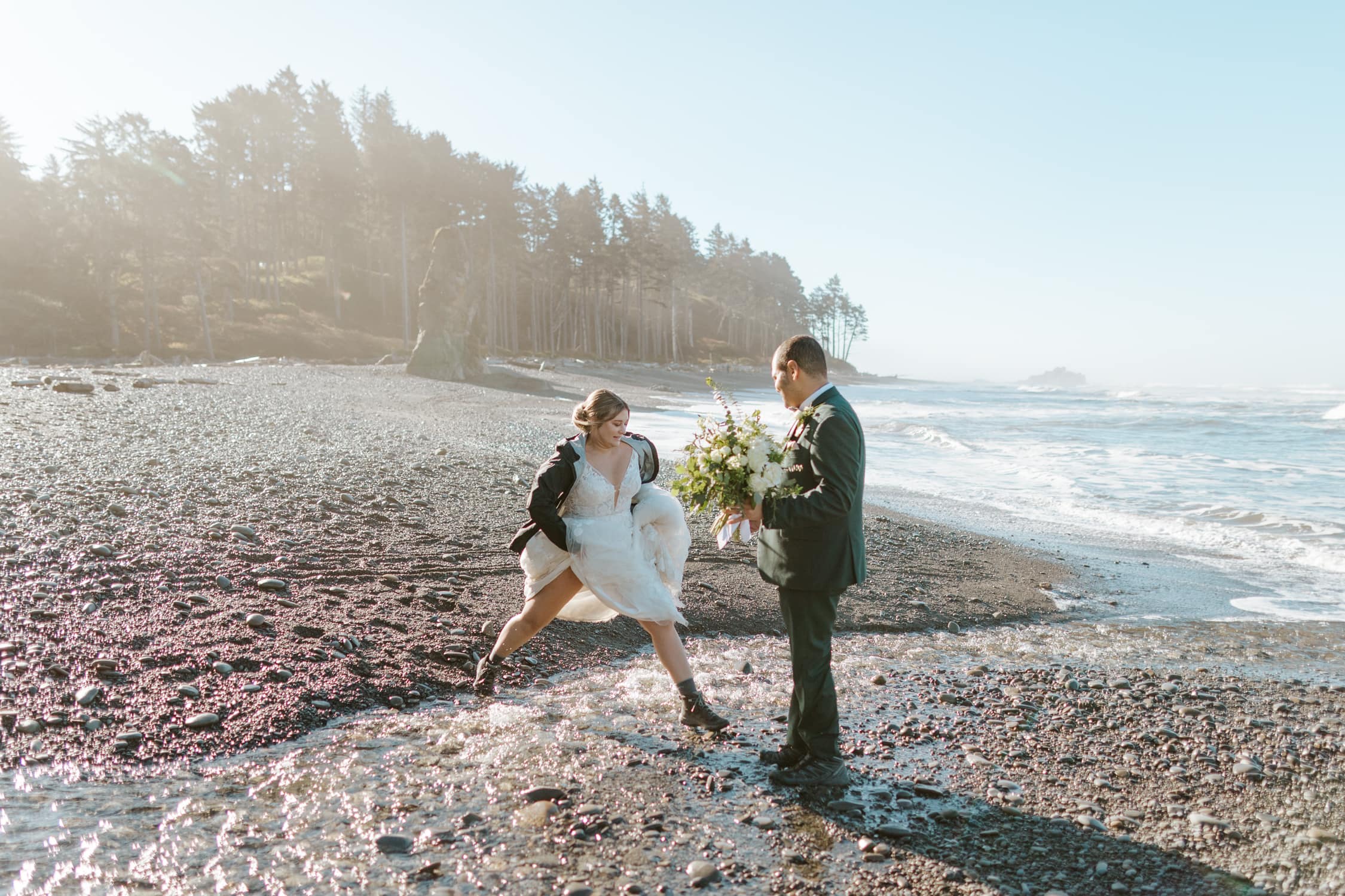 A bride hopping over a stream of water on Ruby Beach while the groom holds her bouquet.