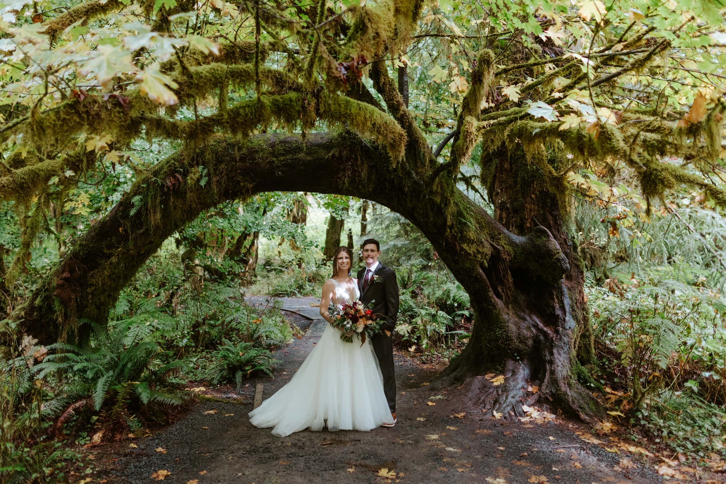 A bride and groom smiling at the camera along the Hall of Mosses Trail in the Hoh Rainforest.