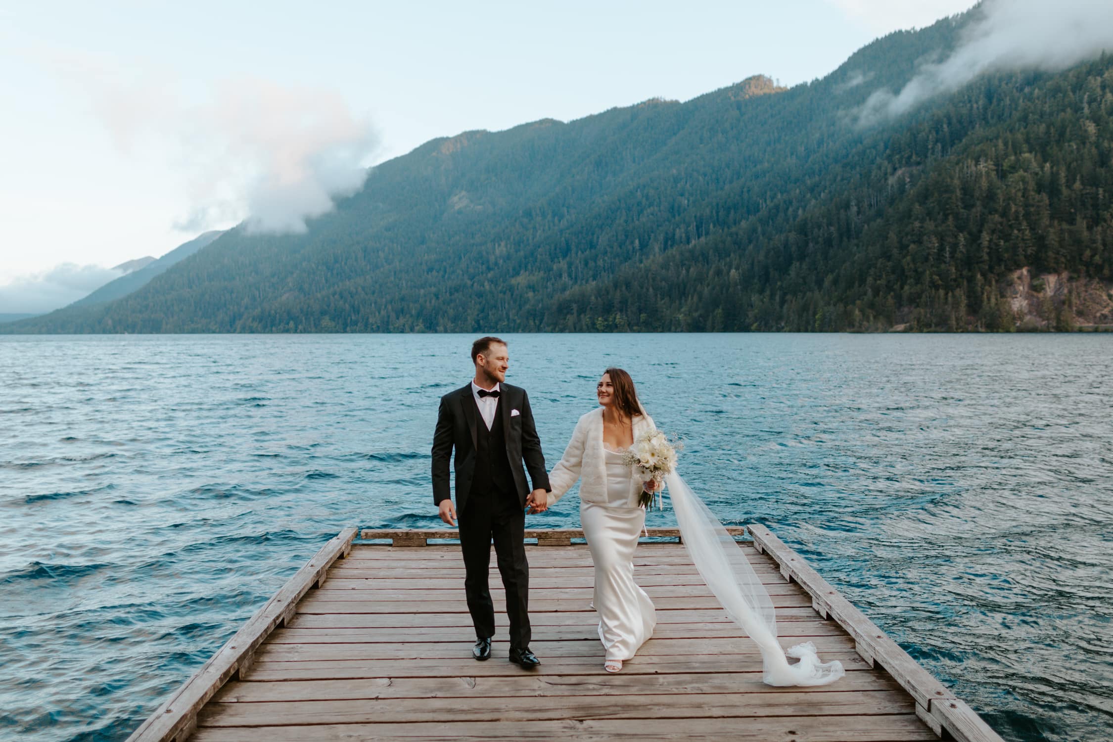 A bride and groom holding hands and walking towards the camera on a dock on Lake Crescent, a blue lake in Olympic National Park.
