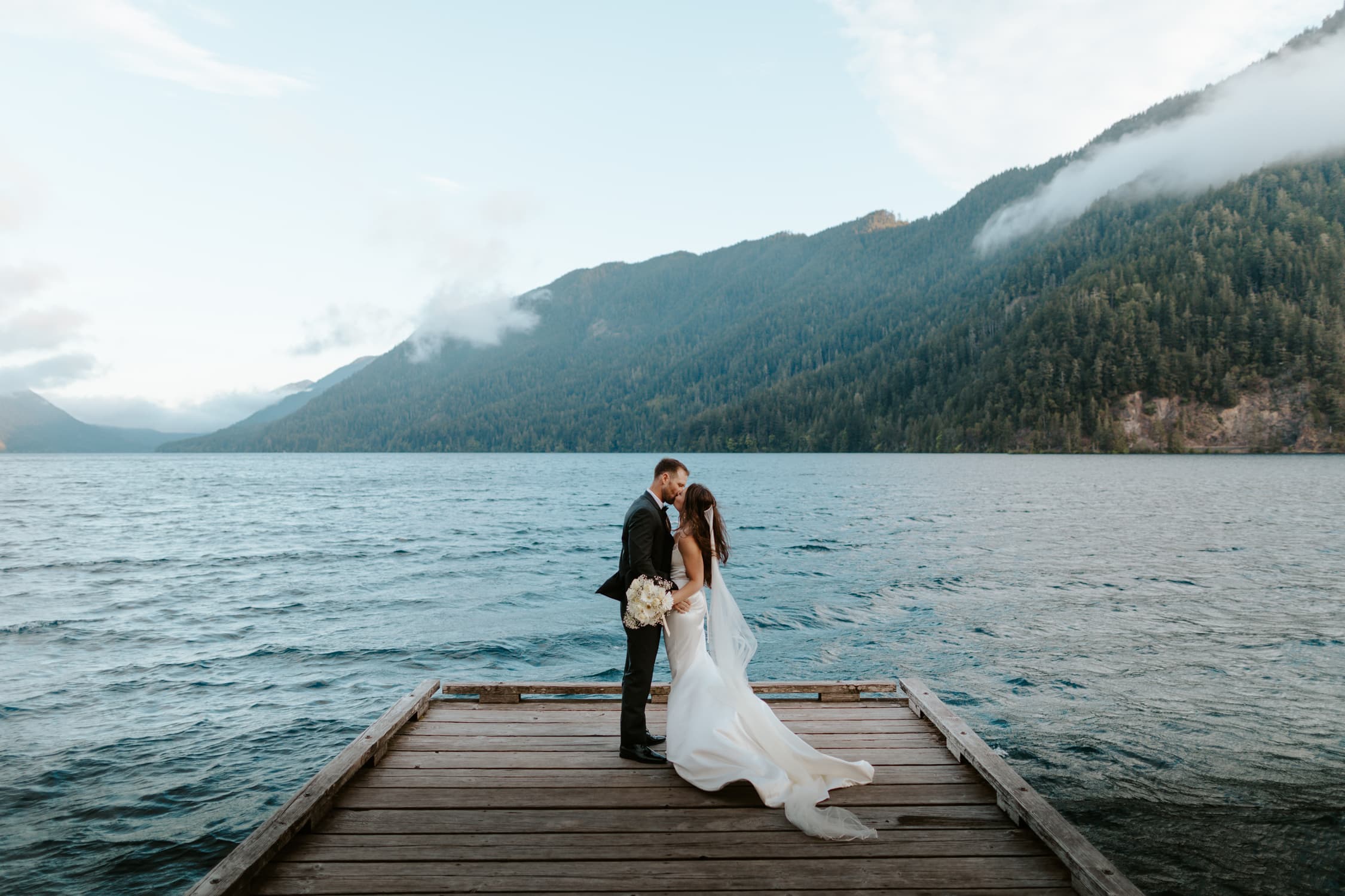 A bride and groom kissing on a dock on Lake Crescent, a blue lake in Olympic National Park.